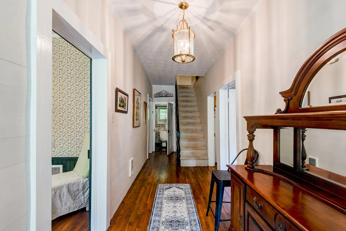 A stylish foyer or transitional interior space with a staircase, console table, chandelier, and art on the walls. The mood is bright and welcoming, giving a sense of arrival and openness.