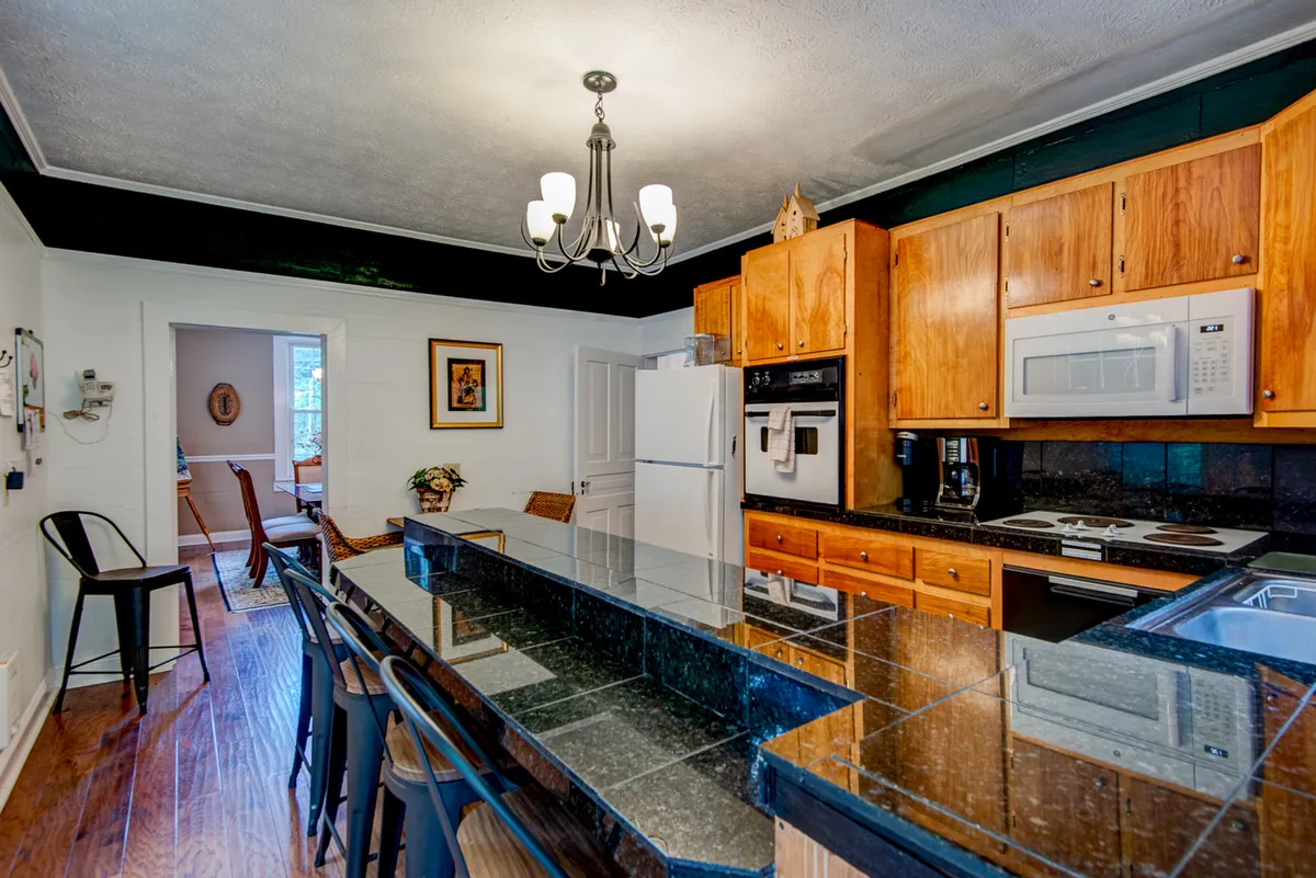 An inviting kitchen scene with a central island topped by sleek black granite, surrounded by wooden cabinets and illuminated by a stylish chandelier. The setup highlights the kitchen's social and practical features, from cooking to entertaining.