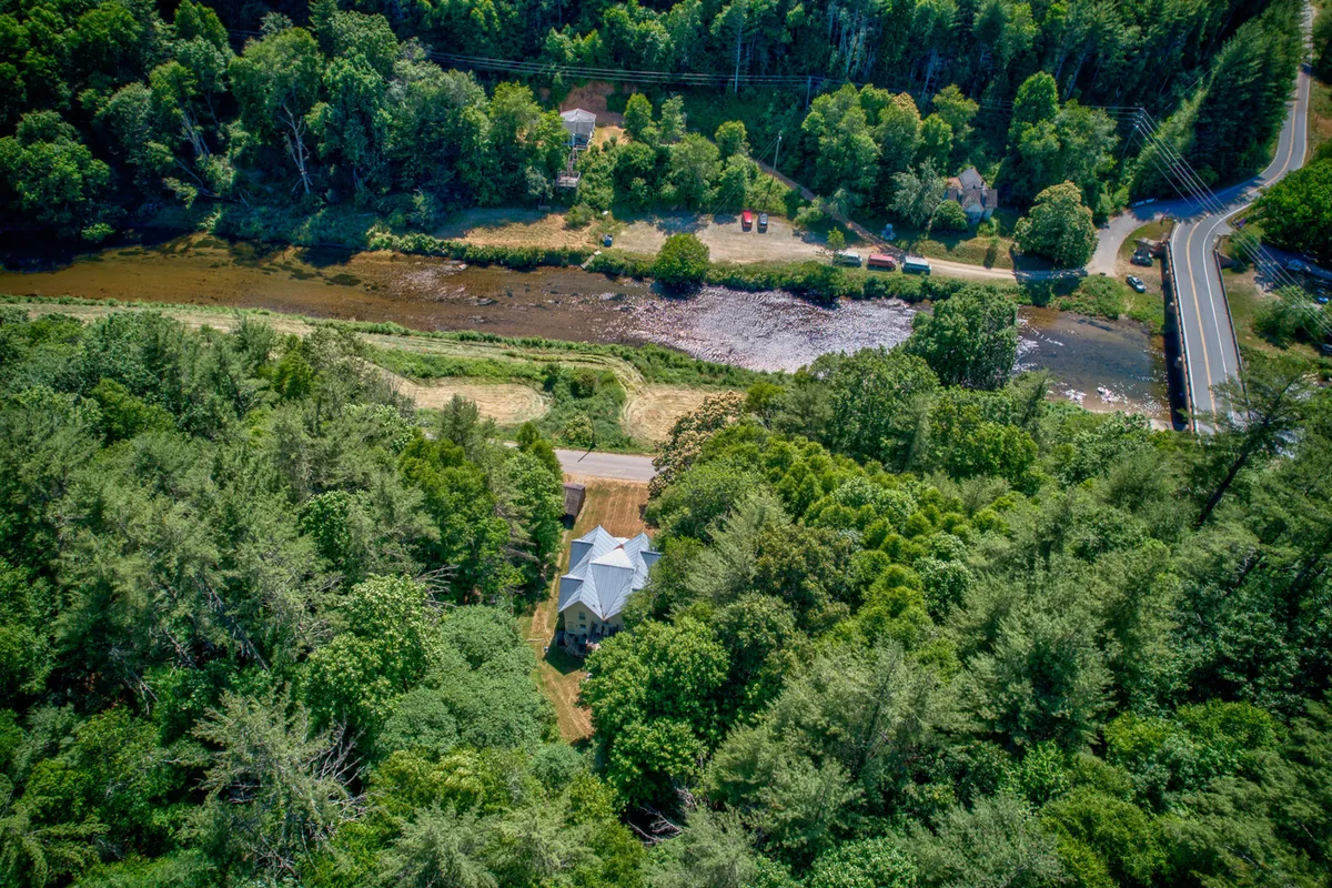 A scenic exterior shot showing the house nestled beside a tranquil river and surrounded by mature trees. The image also captures the road leading to the property, creating a sense of arrival and adventure. The riverbank is visible, hinting at opportunities for fishing, swimming, and tubing.