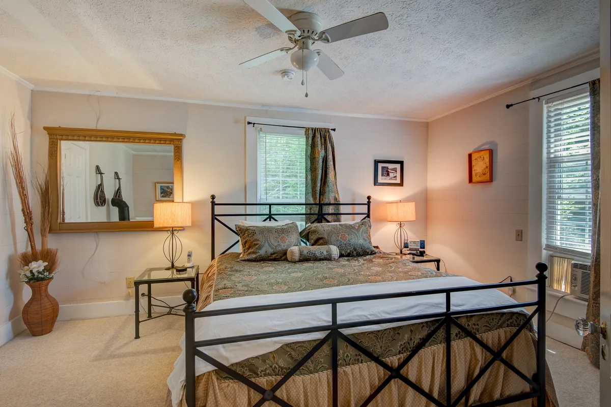 A serene bedroom featuring a neatly made bed with decorative pillows, a framed mirror, table lamps on side tables, and a window with elegant curtains that lets in abundant natural light. The overall palette is soft and the mood is calm and restful.