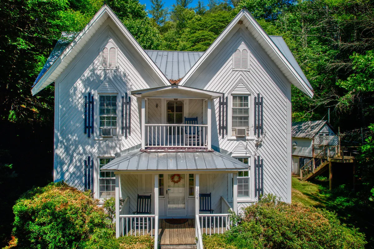 This image showcases the exterior of a charming, historic single-family house with a distinctive A-frame roof, nestled amidst lush greenery. The inviting front porch features classic rocking chairs, perfect for relaxing and soaking in the tranquil river and mountain views. The spacious yard hints at opportunities for gatherings, with the backdrop of trees and natural beauty enhancing the setting's appeal.