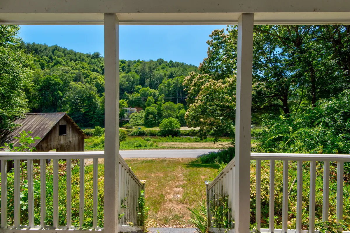 A peaceful porch scene overlooking lush greenery, a distant rustic shed, a meandering road, and gentle hills. The inviting front porch, framed by nature, provides a vantage point for relaxing and observing wildlife or watching the sunrise, perfectly illustrating the restful side of riverfront living.
