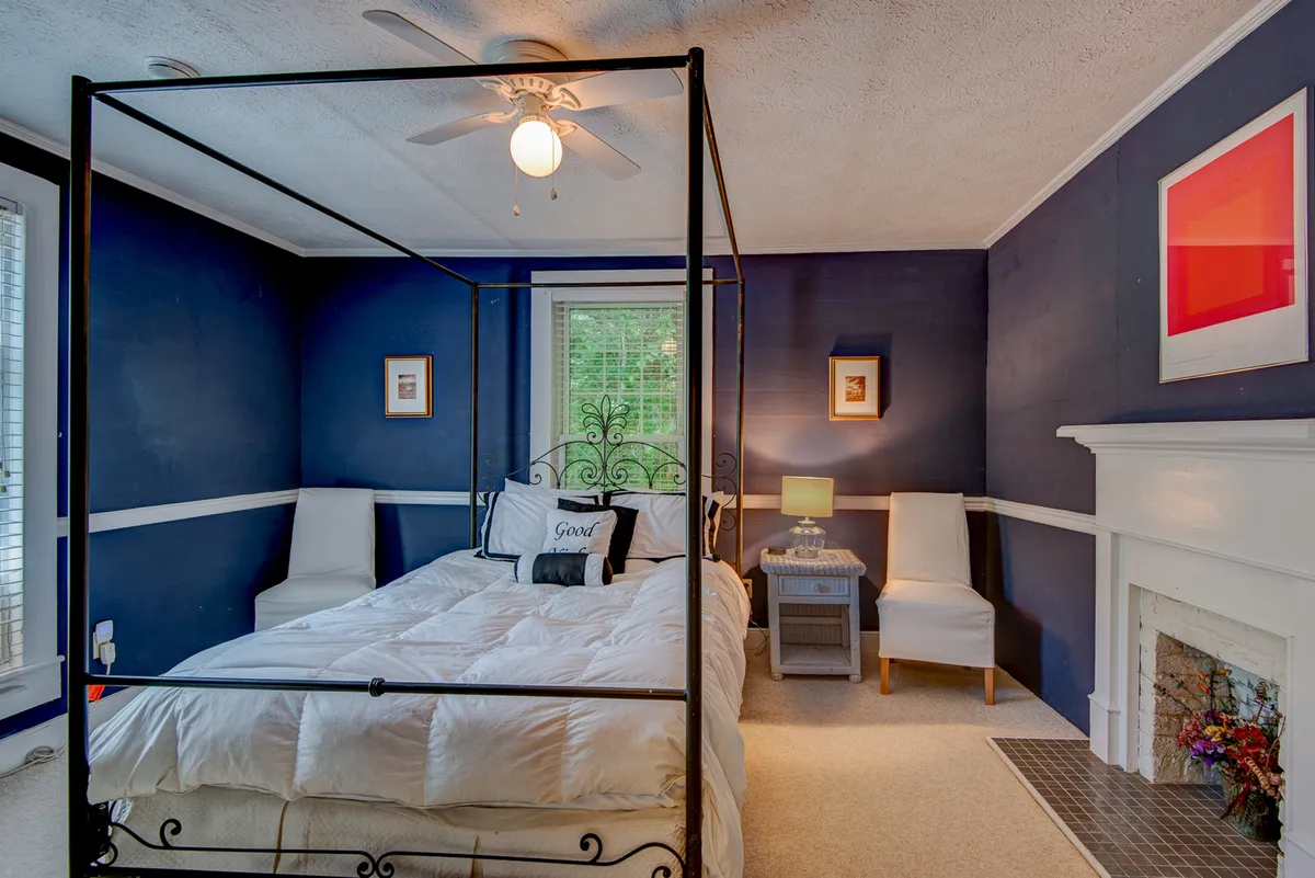 A cozy bedroom with a classic four-poster bed, soft bedding, a fireplace, and thoughtful wall art. A lamp on the nightstand casts a gentle, warm glow, highlighting the room’s calm and literary-inspired ambiance.