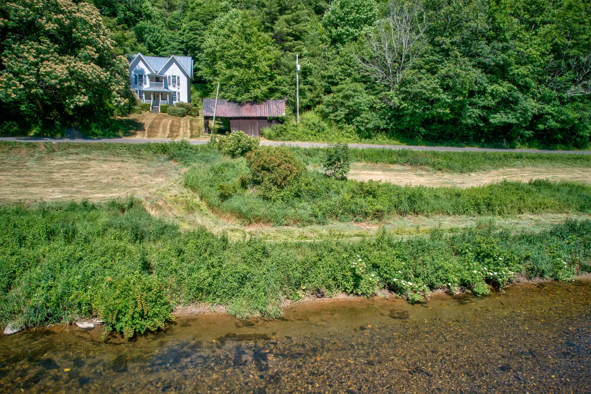 A serene exterior scene featuring the house set beside a beautiful river, with a rustic barn and towering trees in view. The tranquil riverbank invites fishing, while the open grassy area is perfect for picnics and exploring. The lush greenery and gentle sloping land evoke the expansive, natural setting described in the section.