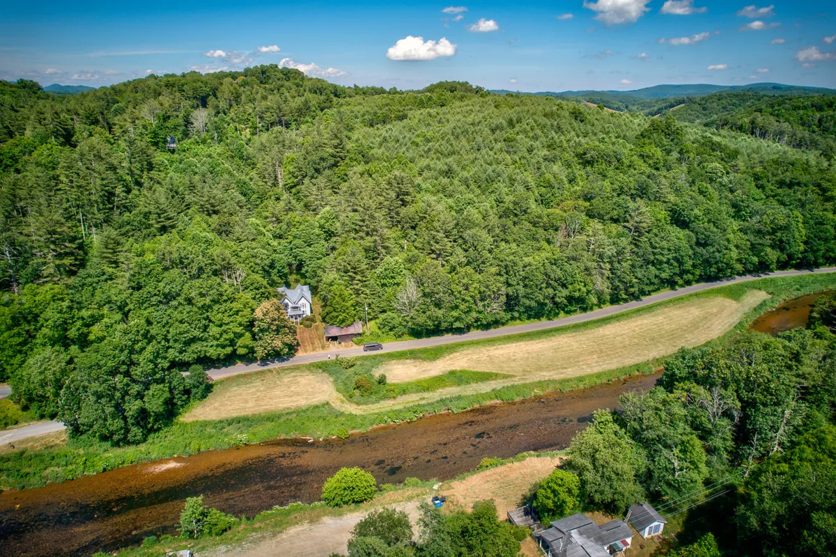 A wide exterior view displaying the house nestled among lush greenery beside a winding river and distant hills. The serene landscape suggests plenty of space for outdoor enjoyment and exploration.