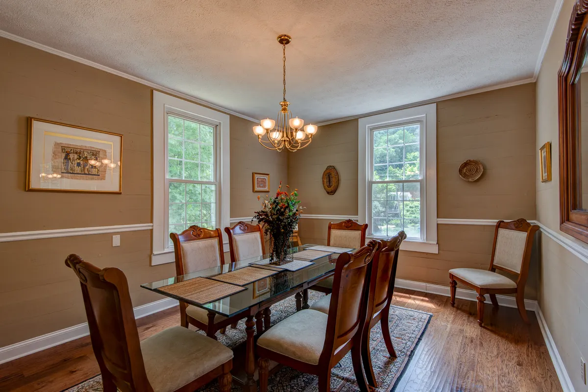 A spacious dining area centered around a glass dining table adorned with a vibrant floral centerpiece. Large windows let in natural light and offer scenic views of the surrounding landscape, while the elegant chandelier above enhances the sense of occasion—ideal for memorable family feasts.