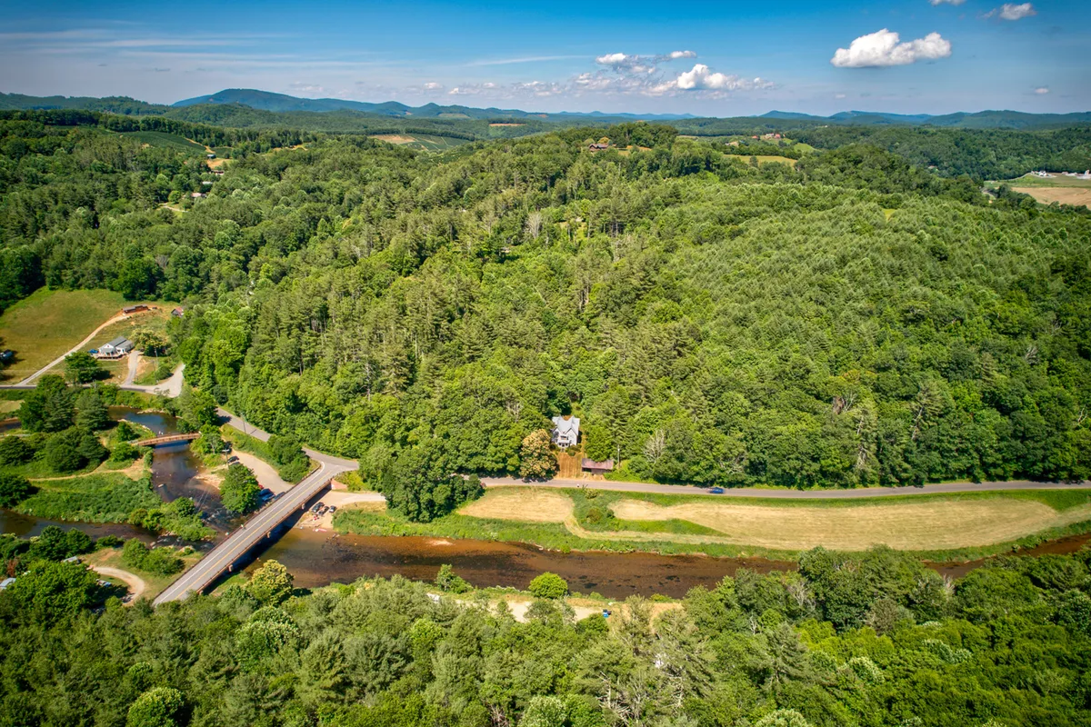 A sweeping view encompassing the river, a wooden bridge, the house, forested areas, and distant mountains. The natural landscape is highlighted by the proximity of water and woodlands, evoking feelings of exploration, wildlife observation, and tranquility. The image hints at multiple activities like hiking, fishing, and nature walks.
