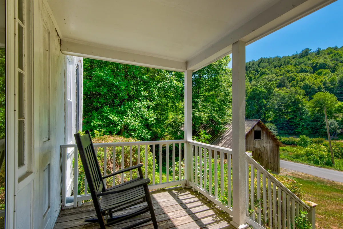 A cozy outdoor setting with a rocking chair on a porch, surrounded by green foliage and a distant barn. The scene feels relaxed and welcoming—perfect for enjoying nature and peaceful moments with pets at your side.