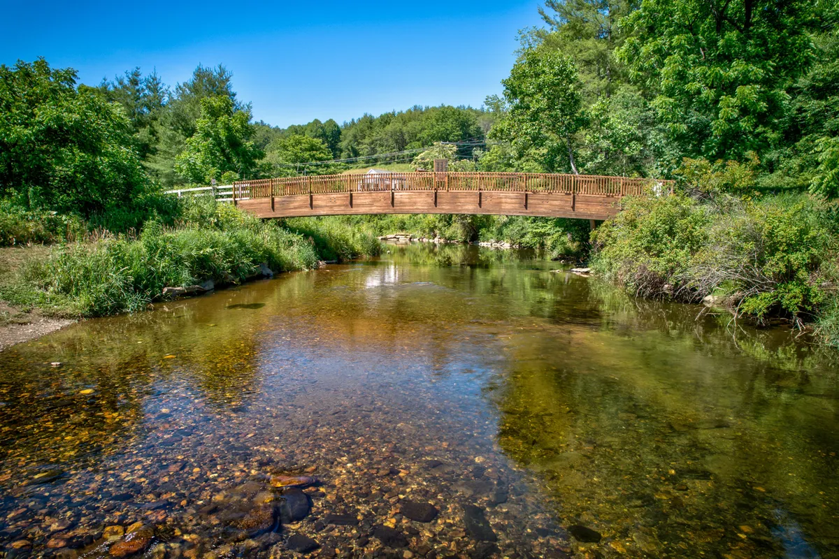 A picturesque scene featuring a wooden bridge over clear water surrounded by lush greenery. The area is peaceful and open, inviting for walks and outdoor activities along the riverbank. Soft sunset reflections add to the tranquil mood.